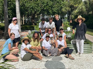 Group of people outdoors sitting and standing on a gravel path with trees and benches in background.