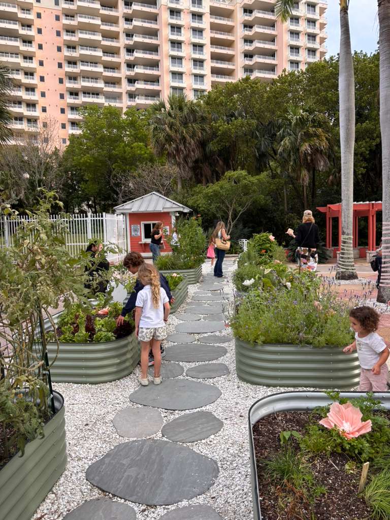 People walking on a garden path with plants and trees, high-rise buildings in the background, and a small red structure nearby.