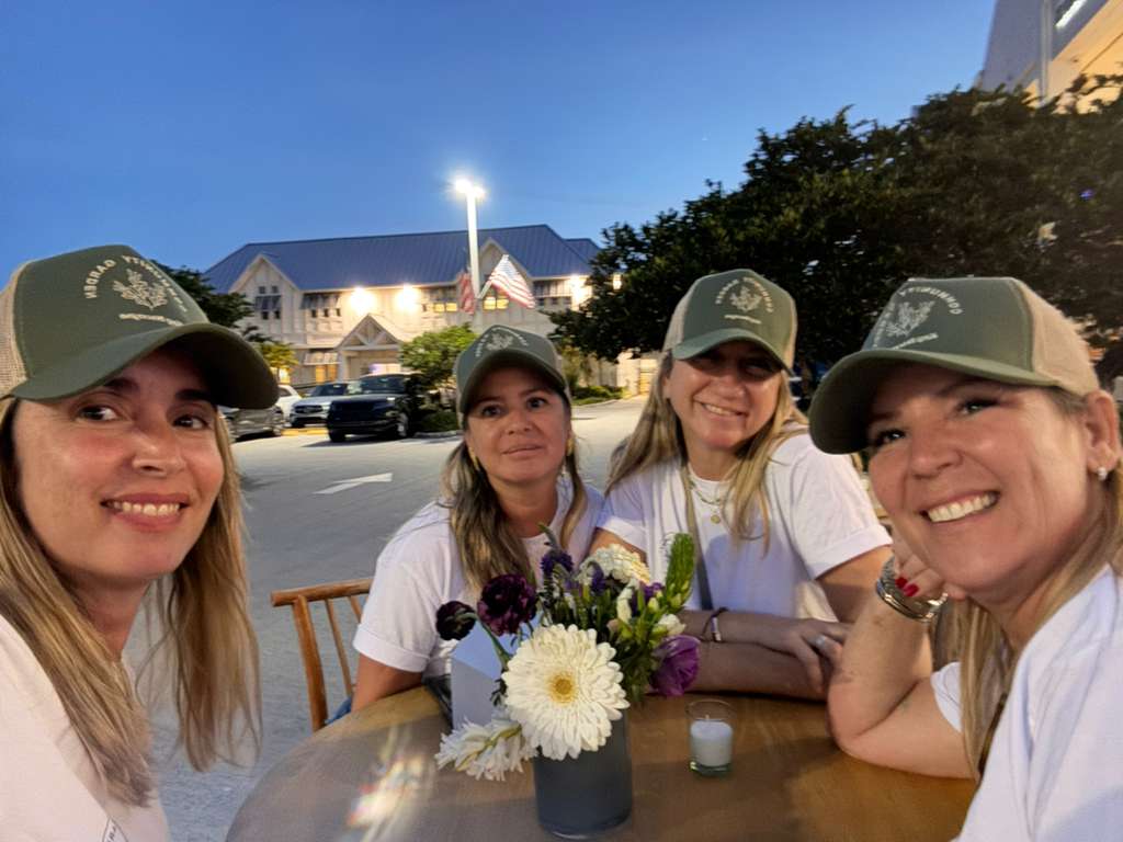 Four women wearing caps and white shirts sit at a table outdoors with a flower arrangement, smiling at the camera.