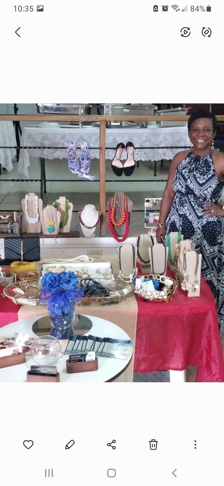 Woman standing next to jewelry display with necklaces, bracelets, and accessories on a table, outdoor setting, sunny day.
