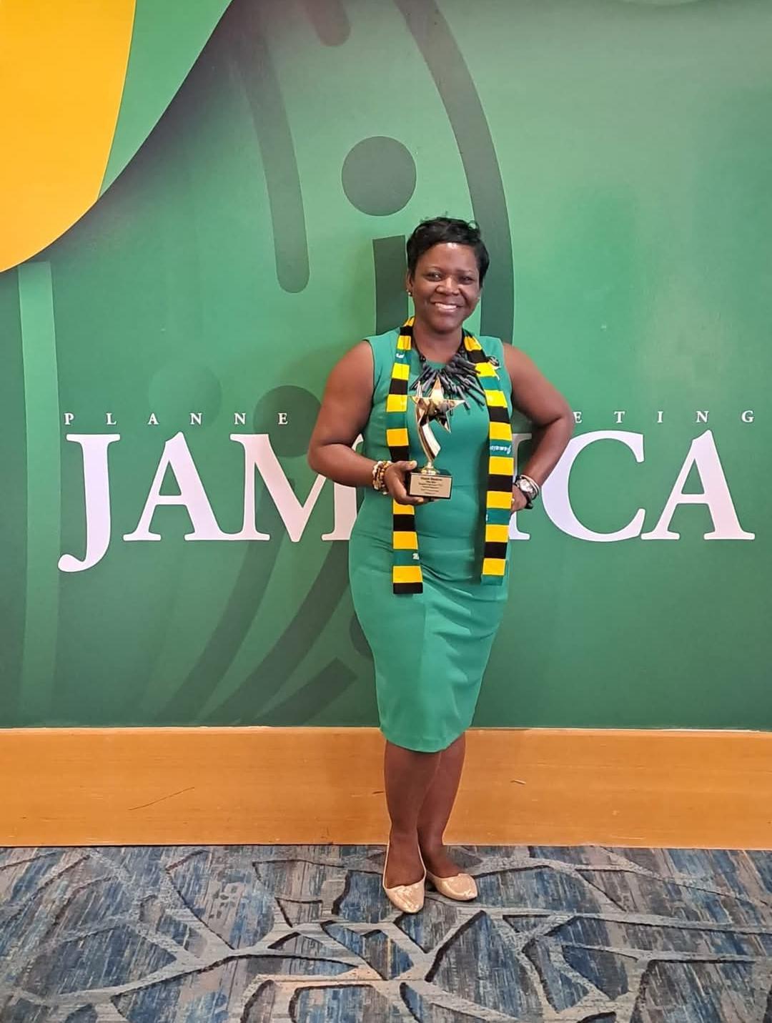 Woman standing in front of a green wall with Jamaica text, wearing a green dress and a colorful scarf, smiling.