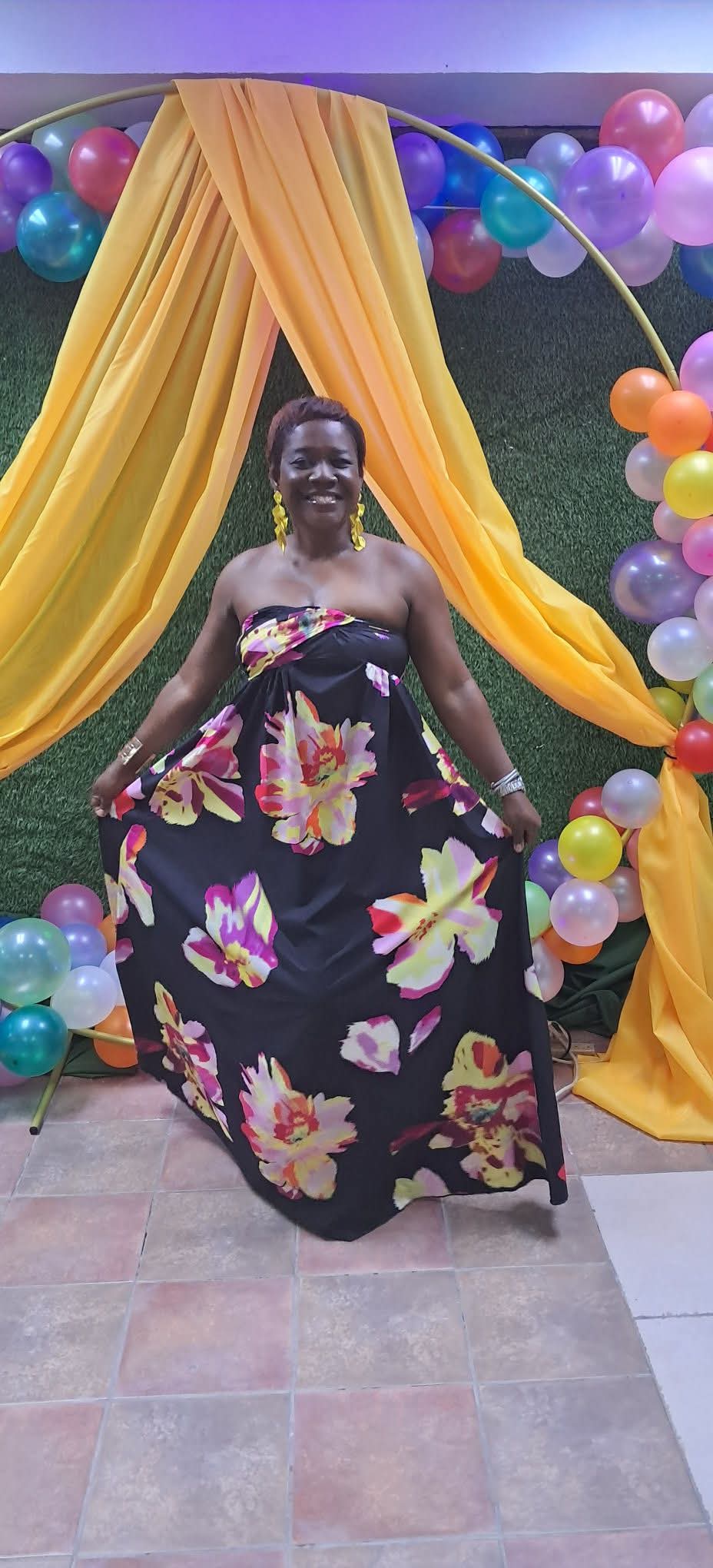 Woman in a floral dress sitting in front of colorful balloons and yellow drapery backdrop.