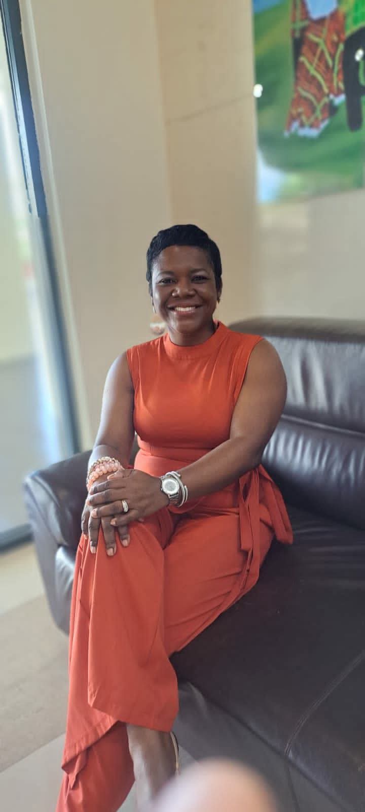 Woman sitting on a black leather couch, smiling, wearing an orange sleeveless dress, with a colorful painting in the background.
