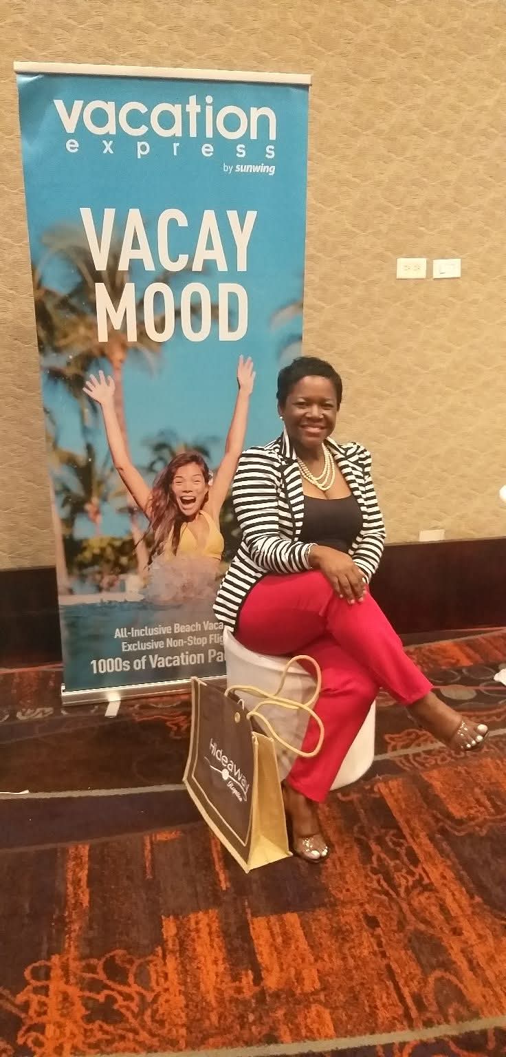 Woman sitting on a stool with a vacation banner behind her, smiling, with a shopping bag on the floor.