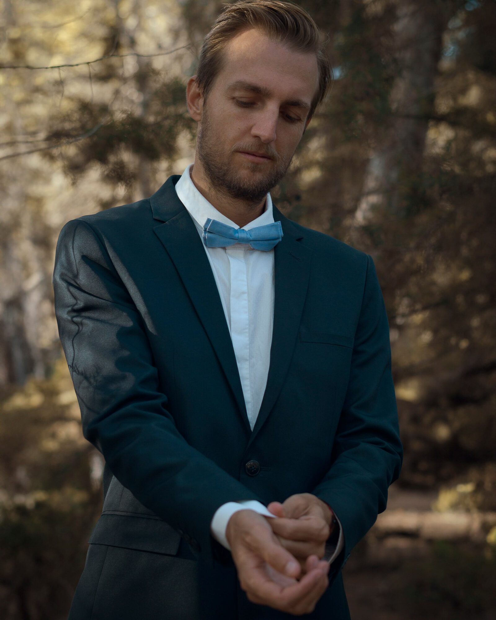 Man in formal suit and bow tie adjusting his cuff in an outdoor setting with trees in the background.