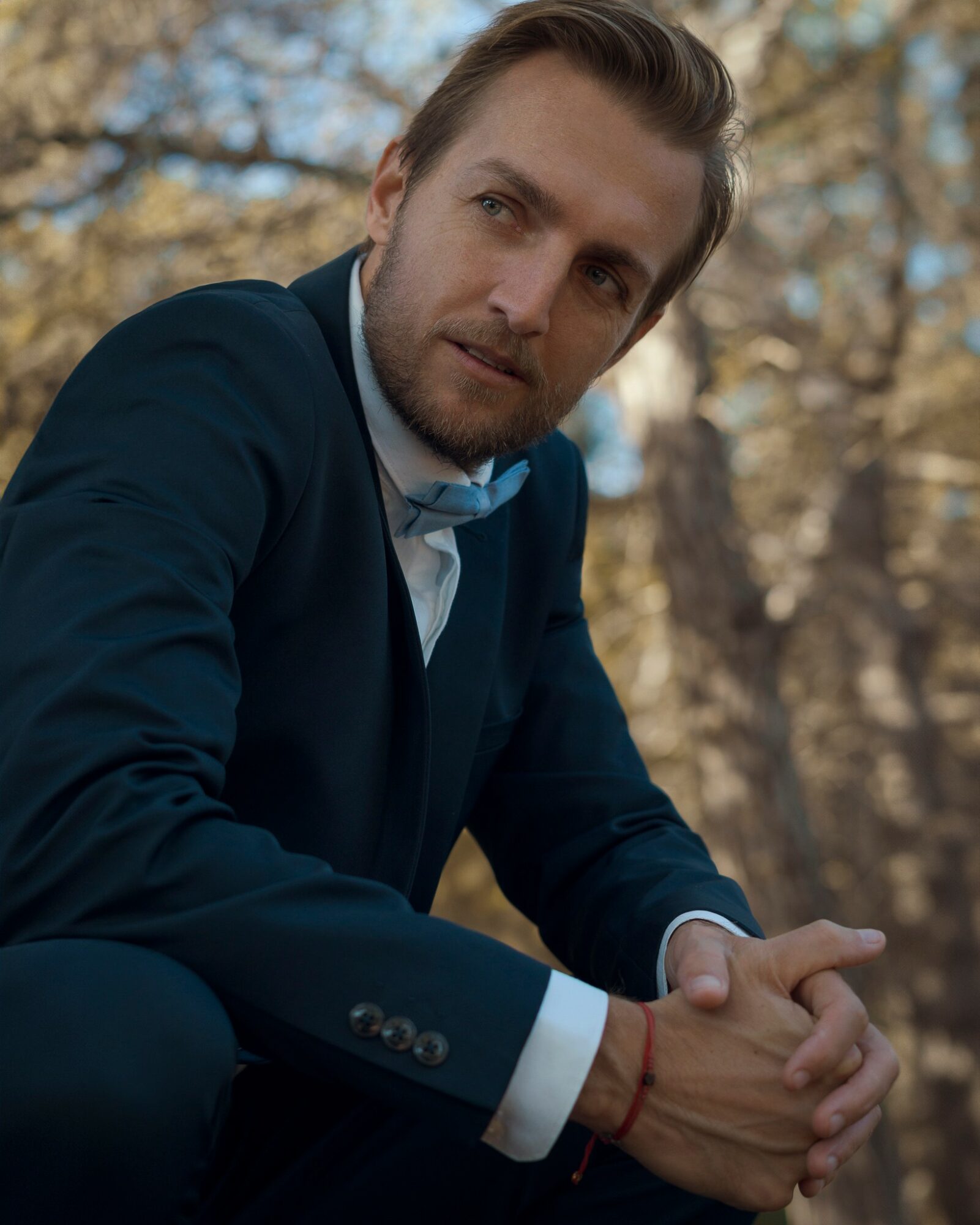 Man in a suit sitting outdoors with trees in the background, looking to the side, hands clasped, sunlight filtering through leaves.