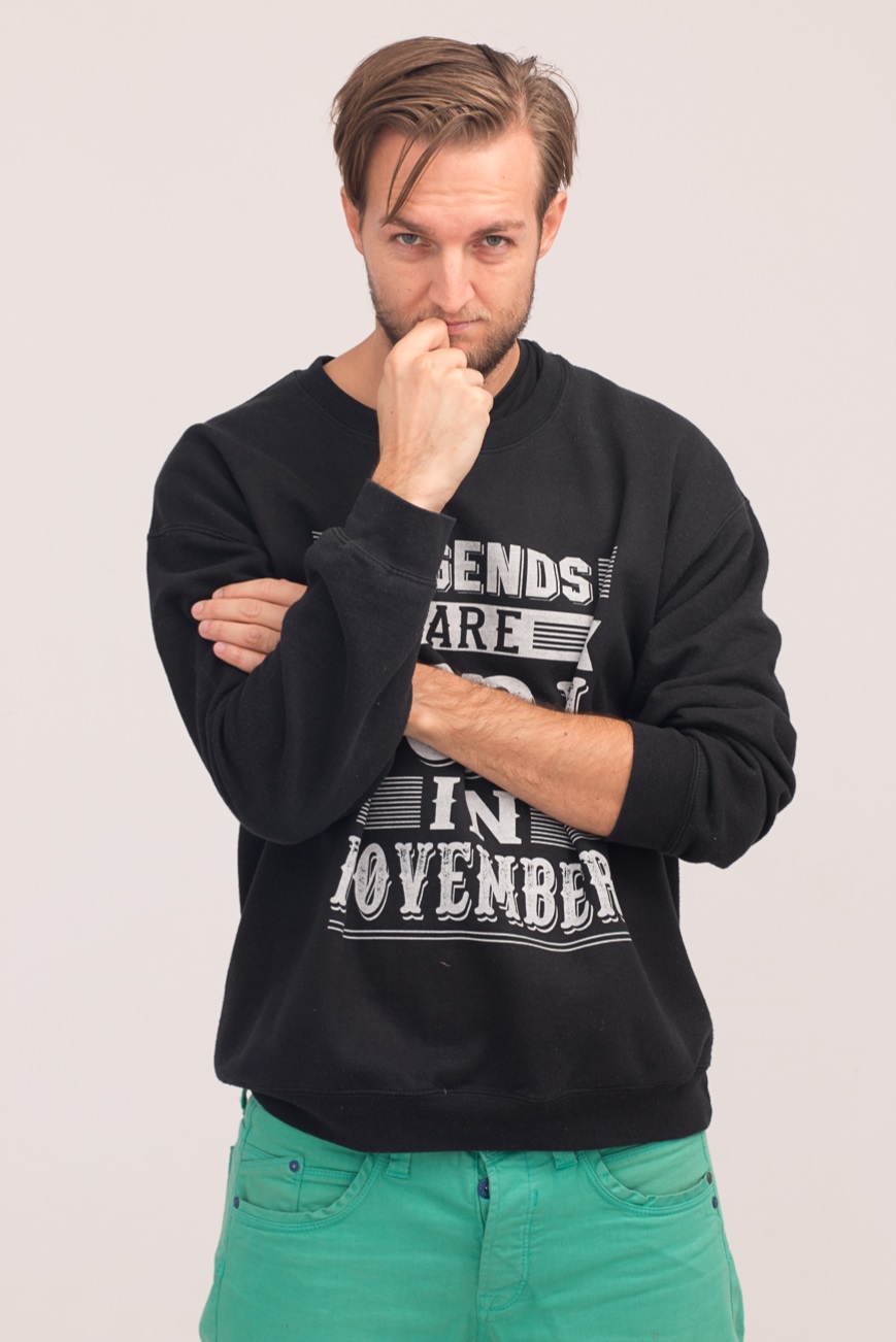 Man with brown hair and beard, wearing a black sweatshirt and green pants, standing with hand on chin, looking thoughtful.