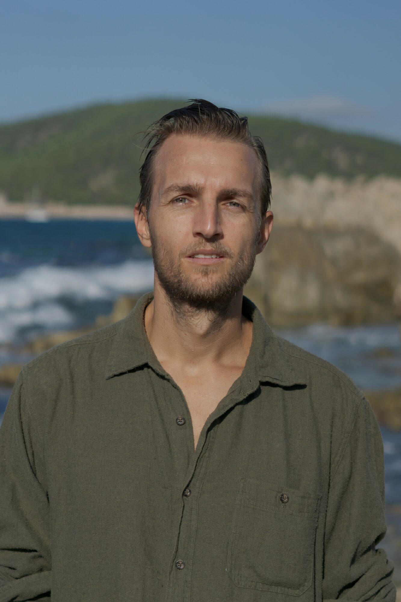 Man with short hair and beard outdoors near water, with a hill in the background, wearing a green shirt.