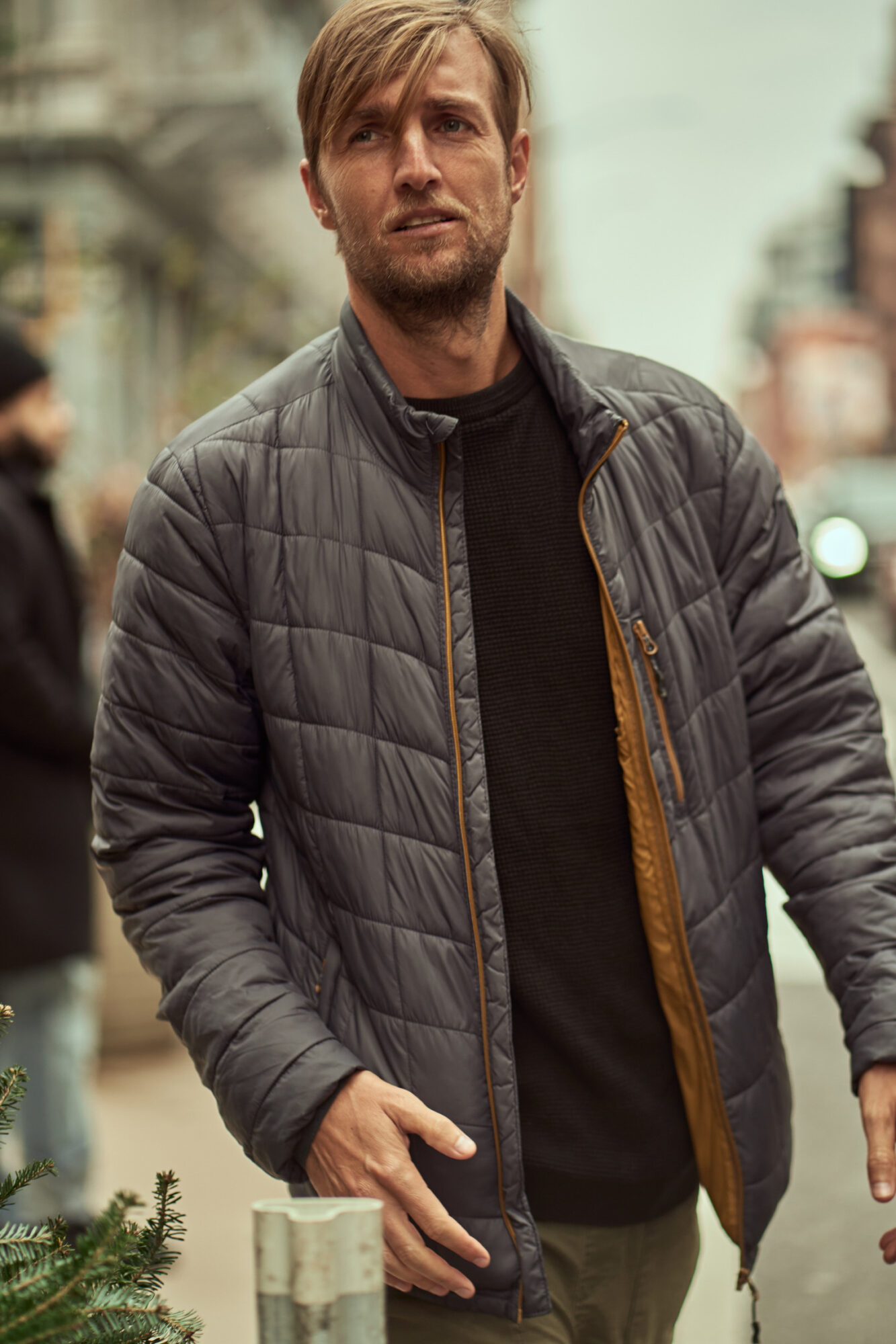 Man with light brown hair and beard wearing a gray quilted jacket and black shirt standing outdoors in an urban setting