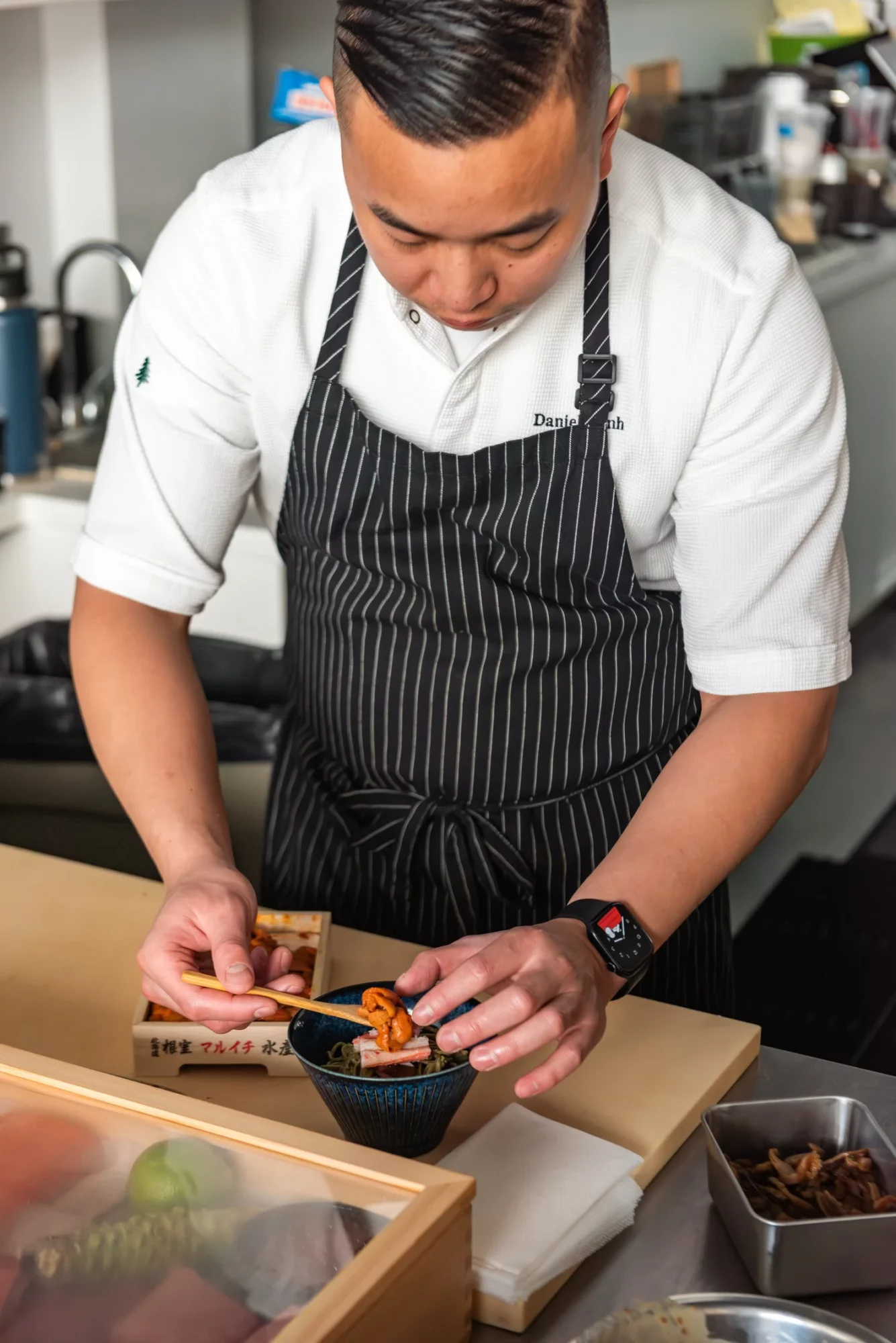 Chef in striped apron preparing food in a kitchen, focused on dish, with various kitchen tools around.