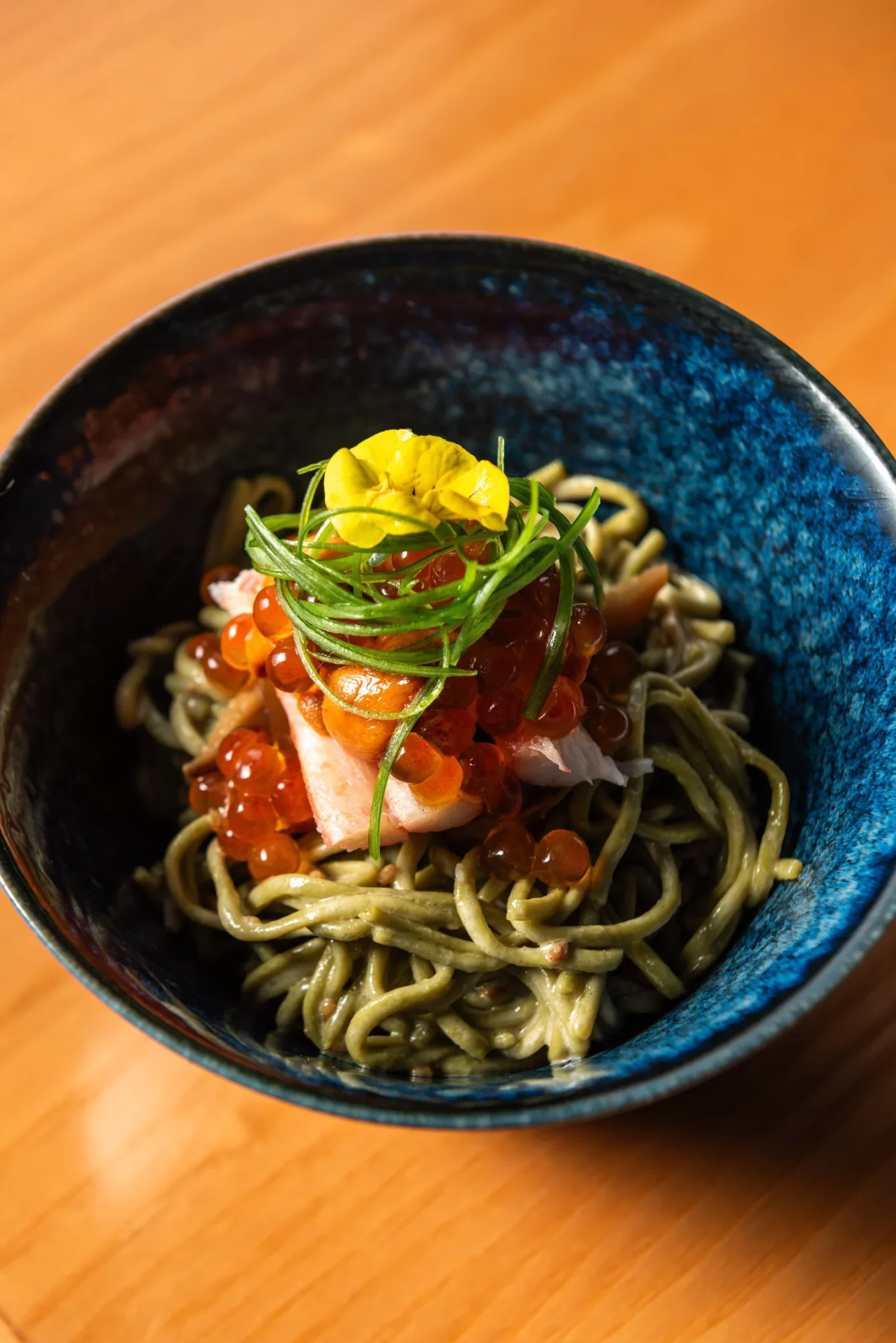 Bowl of pasta with cherry tomatoes, green herbs, and yellow edible flower on top, on a wooden surface.