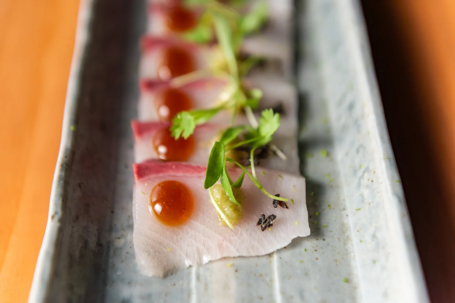 Thin slices of fish topped with green herbs and small orange spheres, arranged on a rectangular plate.