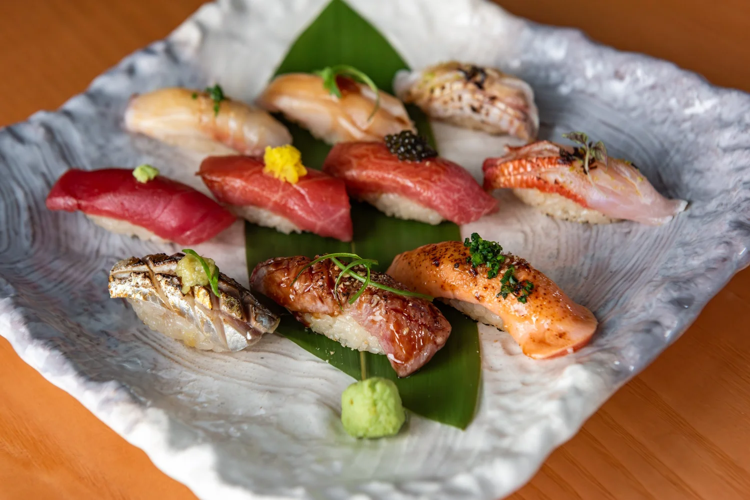 Assorted sushi pieces arranged on a silver plate with a green leaf, wasabi, and garnishes, on a wooden surface.