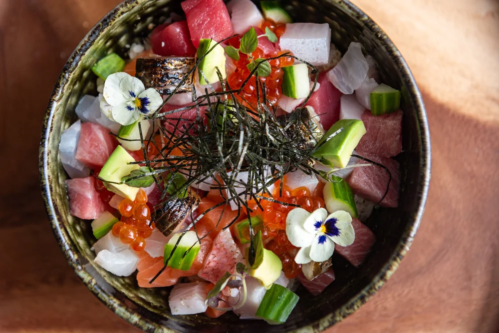 Bowl of diced raw fish, vegetables, and garnishes with edible flowers and seaweed, arranged in a colorful presentation.