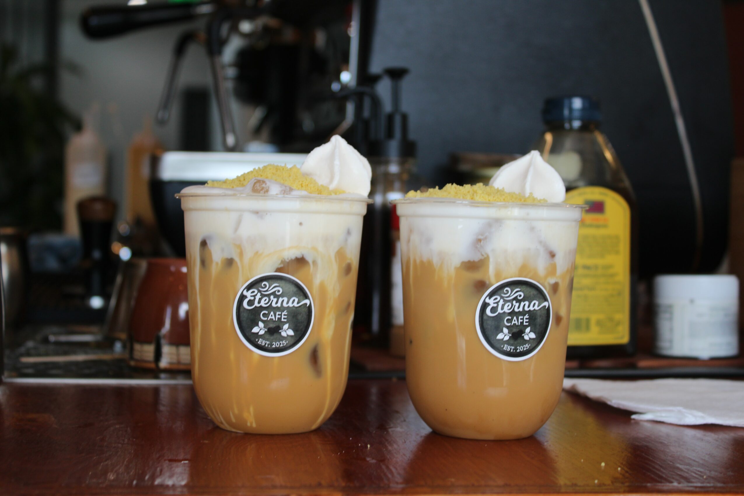 Two iced coffee drinks topped with whipped cream and lemon slices on a bar counter, with kitchen items in background.