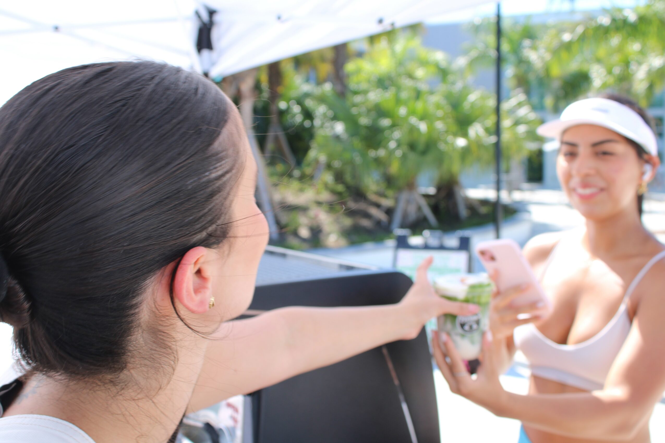 Two women taking a selfie outdoors, one holding a phone, smiling, with greenery in the background.