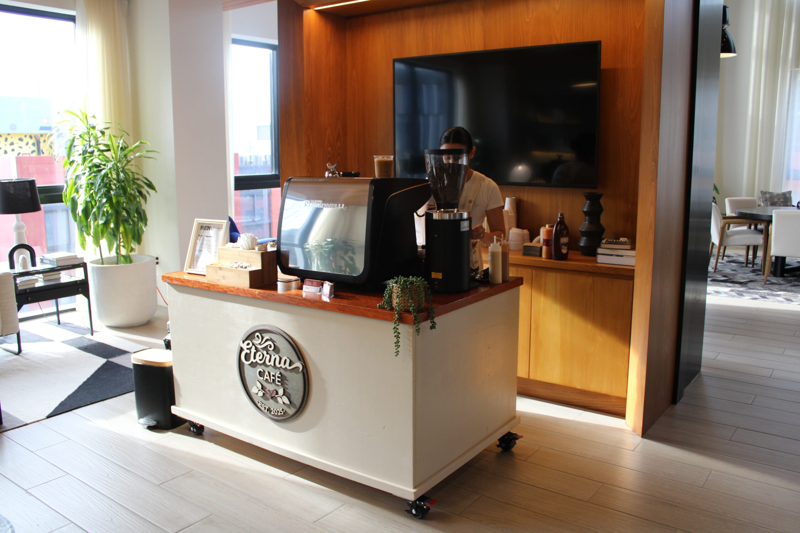 Coffee station with a white counter, coffee machine, and a large TV behind it in a bright room with windows and plants.