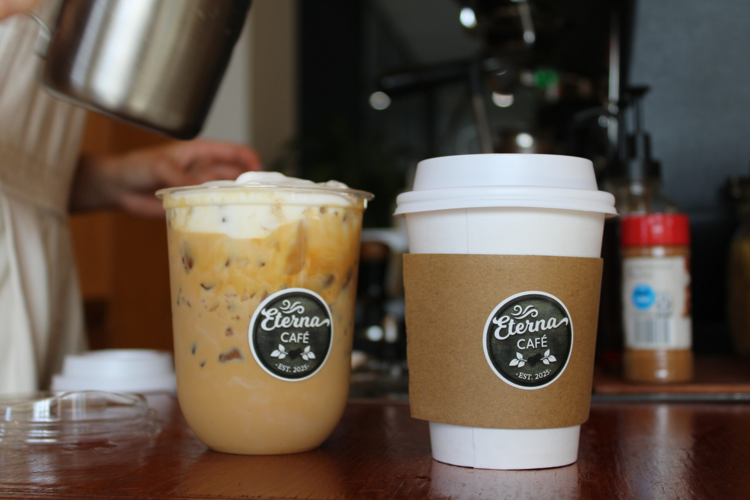 Two cups on a wooden table, one with a cold beverage and the other a hot drink, in a cafe setting.