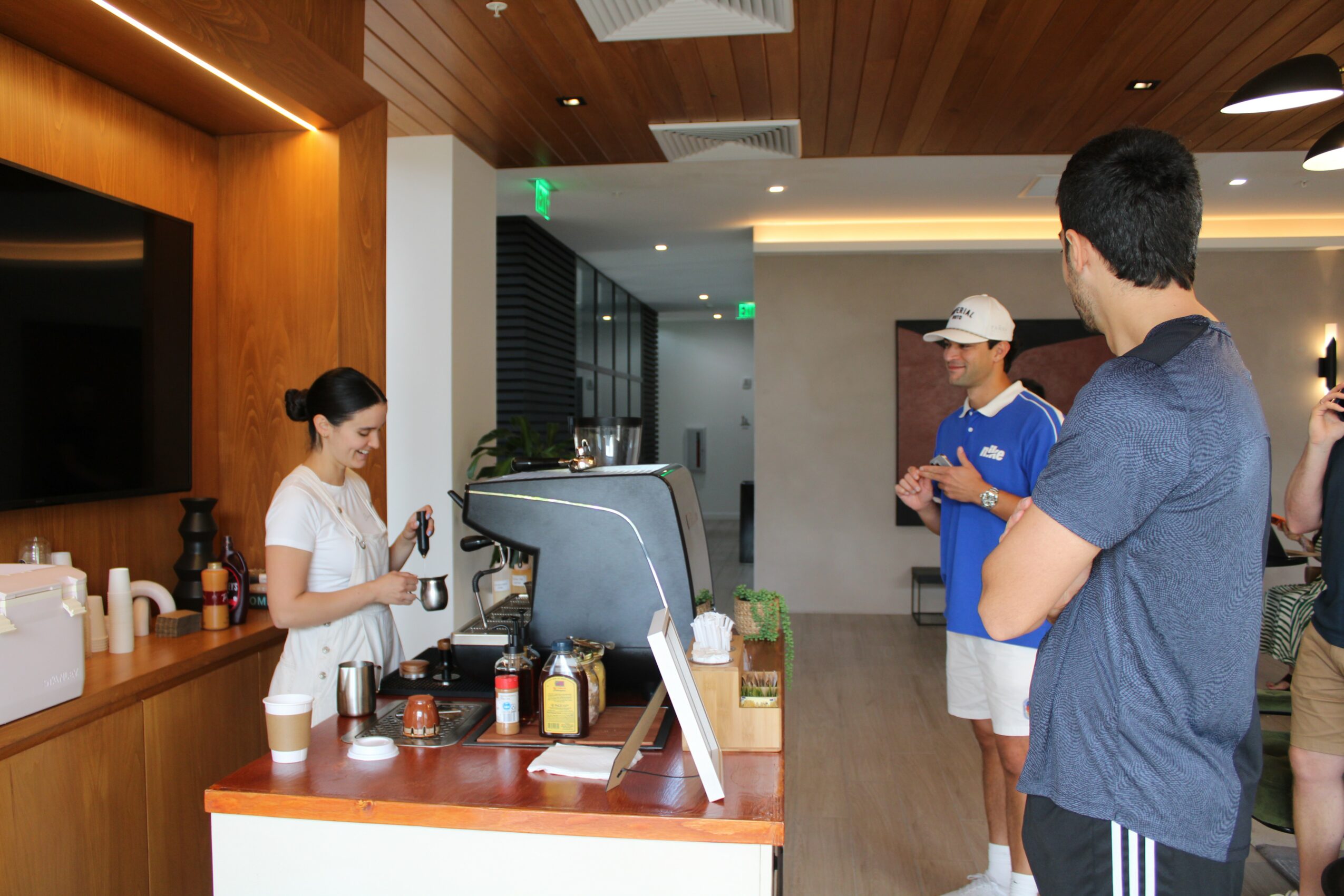 People in a cafe or restaurant, one person preparing drinks, others waiting, modern interior with wooden ceiling and lighting.