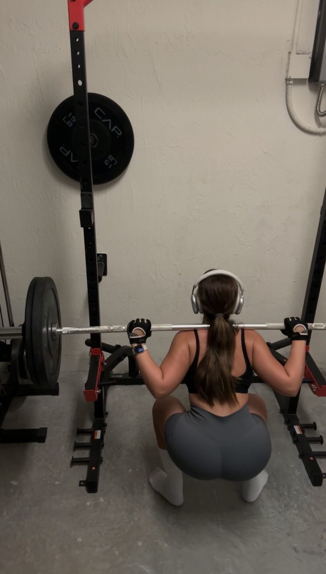 Woman with long hair in a ponytail, wearing headphones, squatting with a barbell on her shoulders in a gym.