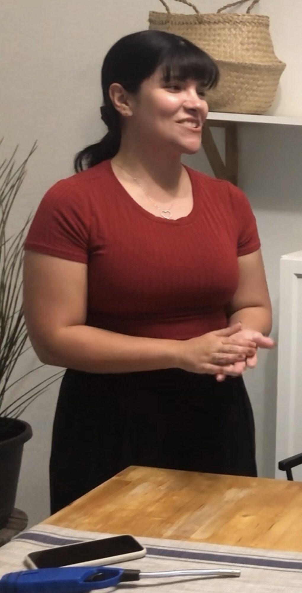Woman with dark hair in a ponytail, wearing a red top, standing indoors near a table and a plant.