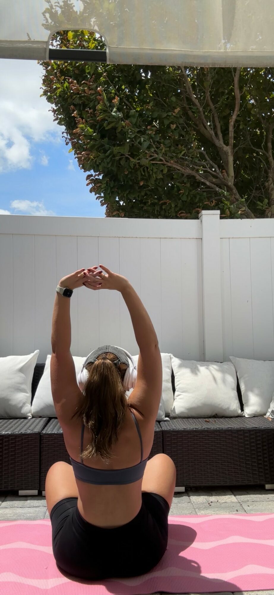 Person sitting on pink mat outdoors, stretching arms overhead, with white fence, sofa, and tree in background.