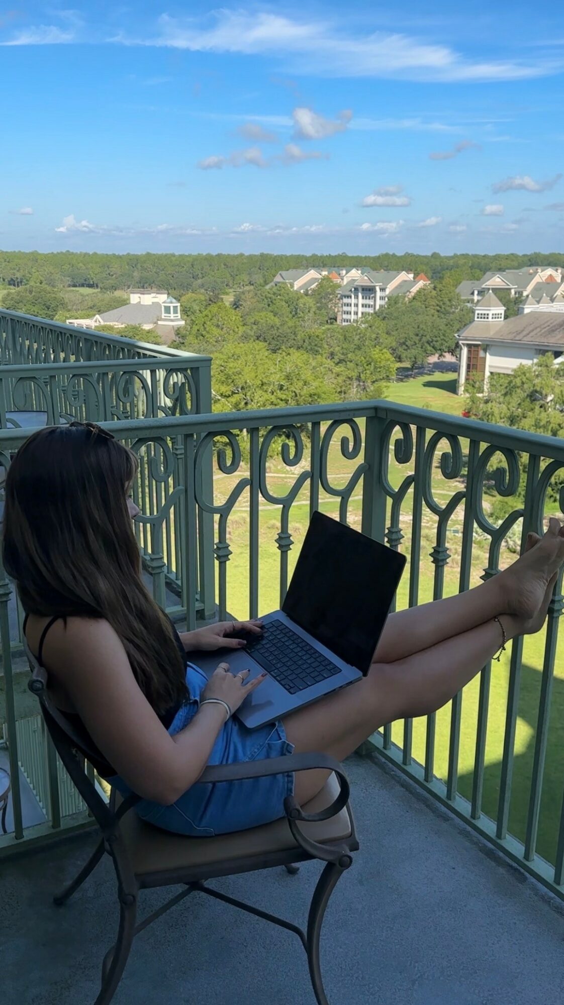 Girl sitting on a balcony using a laptop, overlooking a green landscape with buildings and a blue sky.