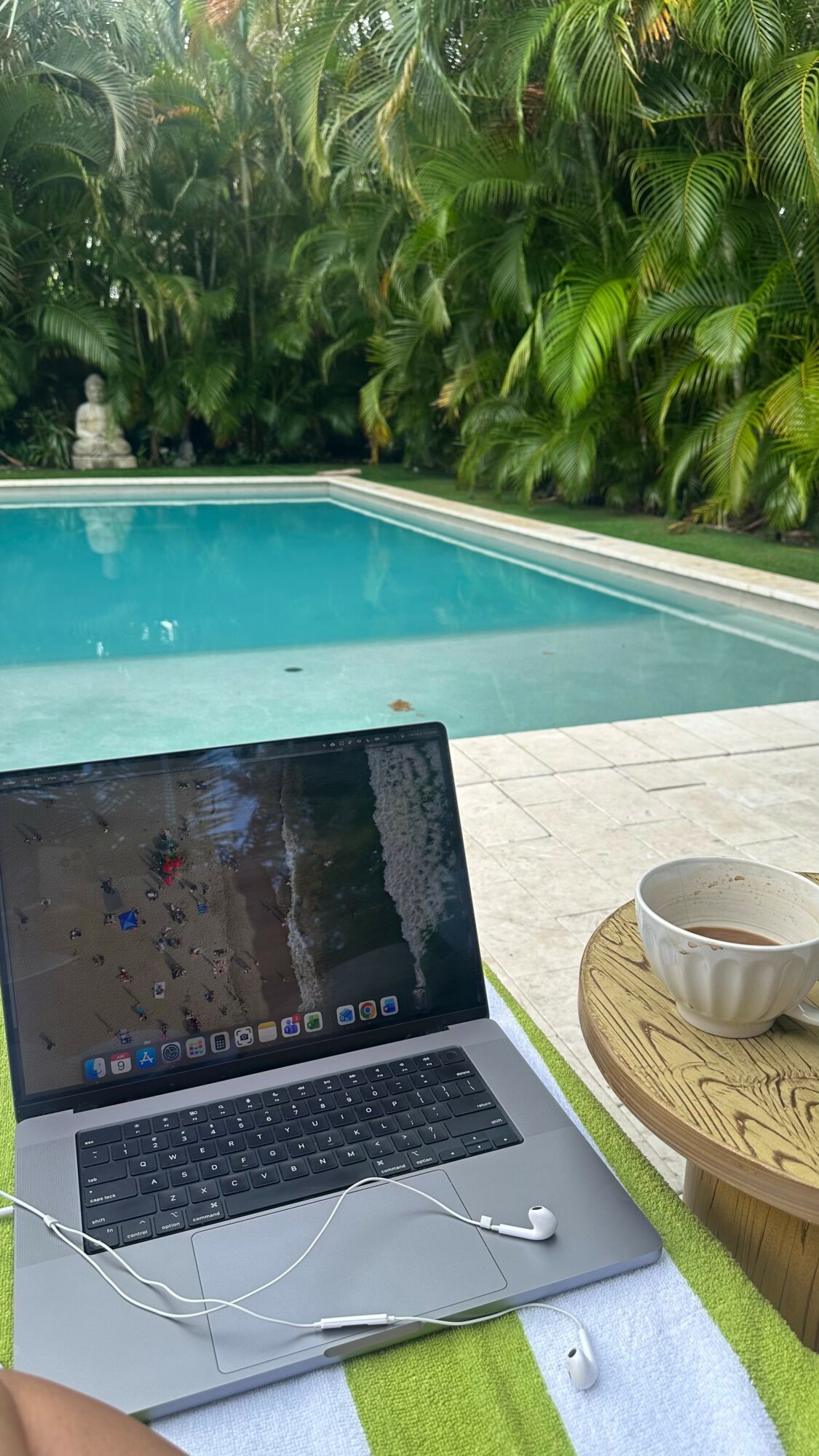 Laptop and bowl on a table near a swimming pool surrounded by lush green plants.