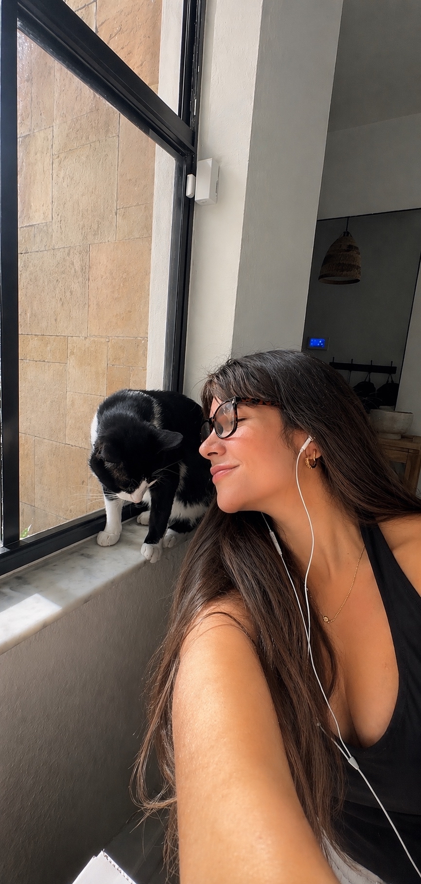 Woman with long hair and glasses leaning near a window, looking at a black and white cat on the windowsill.