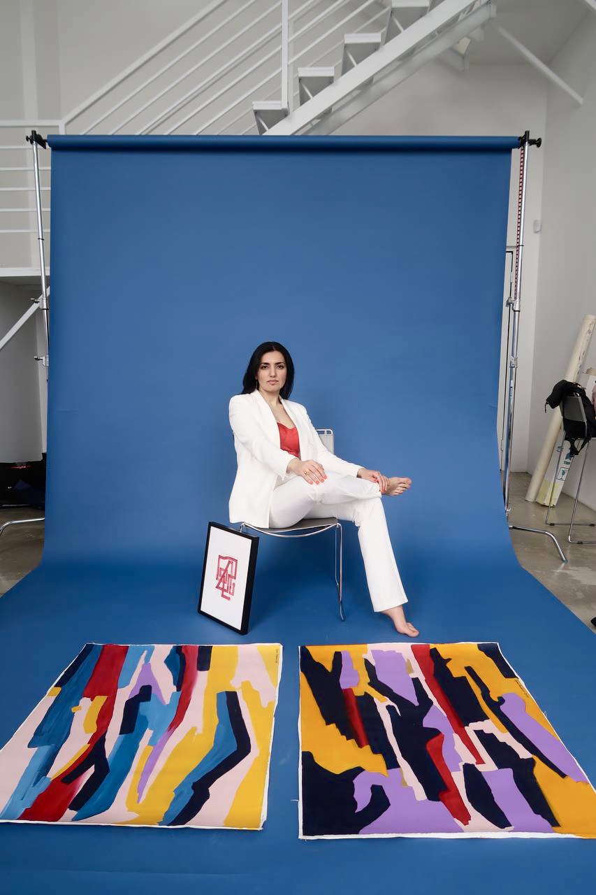 Woman in white suit sitting on a chair in front of a blue backdrop, with colorful abstract rugs on the floor.