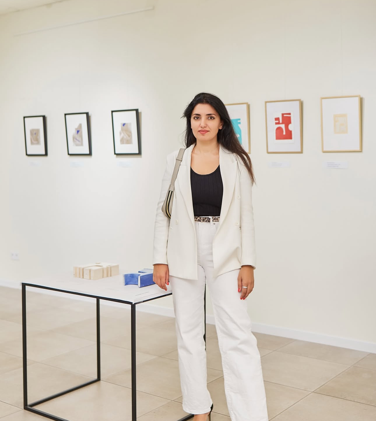 Woman in white suit standing in art gallery with framed artwork on white wall behind her.