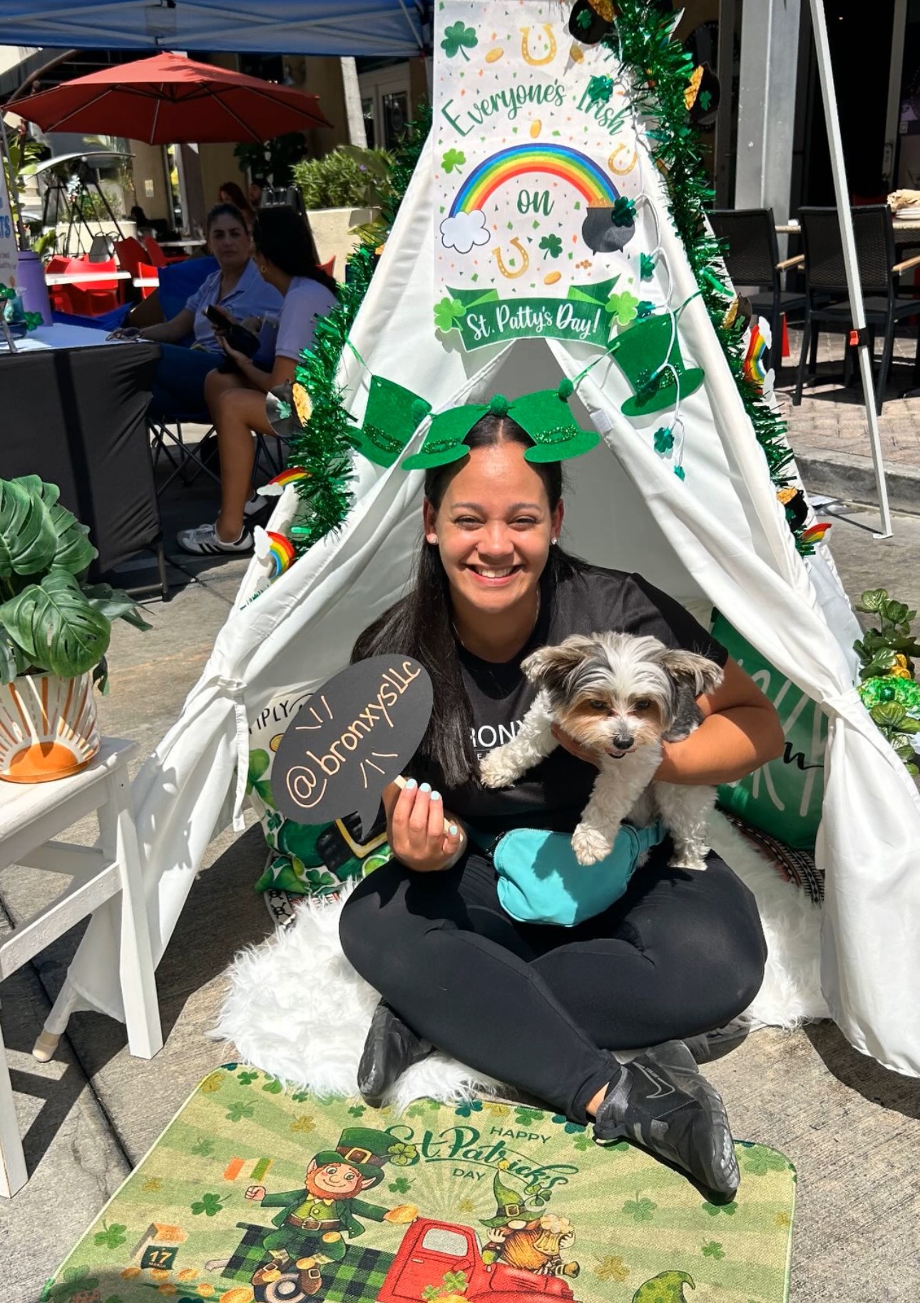Person sitting inside a decorated teepee tent holding a small dog, smiling, with outdoor tables and people in the background.