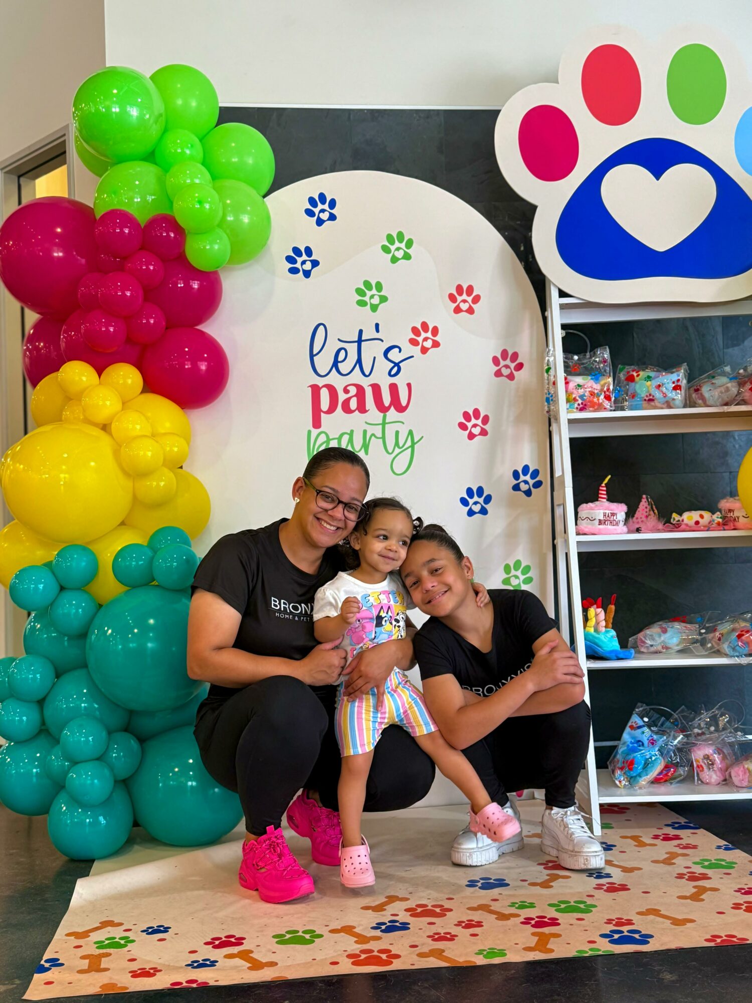 Two women and a girl pose in front of a paw print backdrop with colorful balloons at a pet-themed party, smiling.