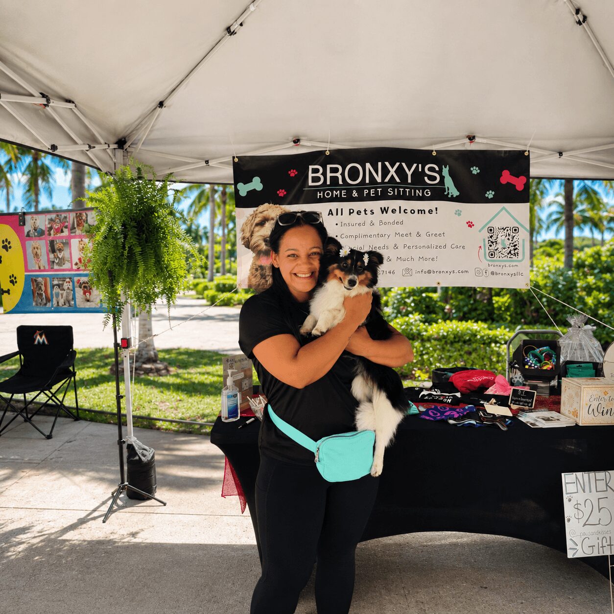 Woman holding a dog under a tent at a pet sitting event, with a banner and tables behind her.