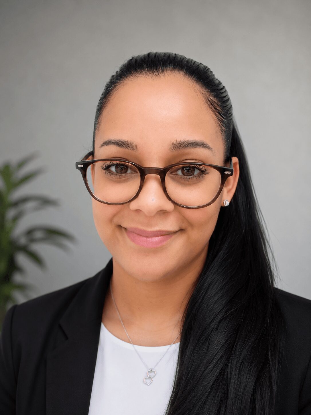 Portrait of a woman with glasses, smiling, wearing a black blazer and white top, with a plant in the background.