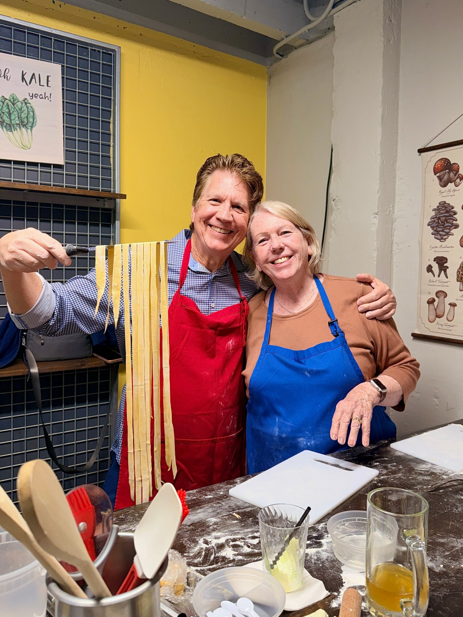 Two smiling women in aprons stand behind a counter with kitchen utensils and glasses, embracing each other in a colorful room.