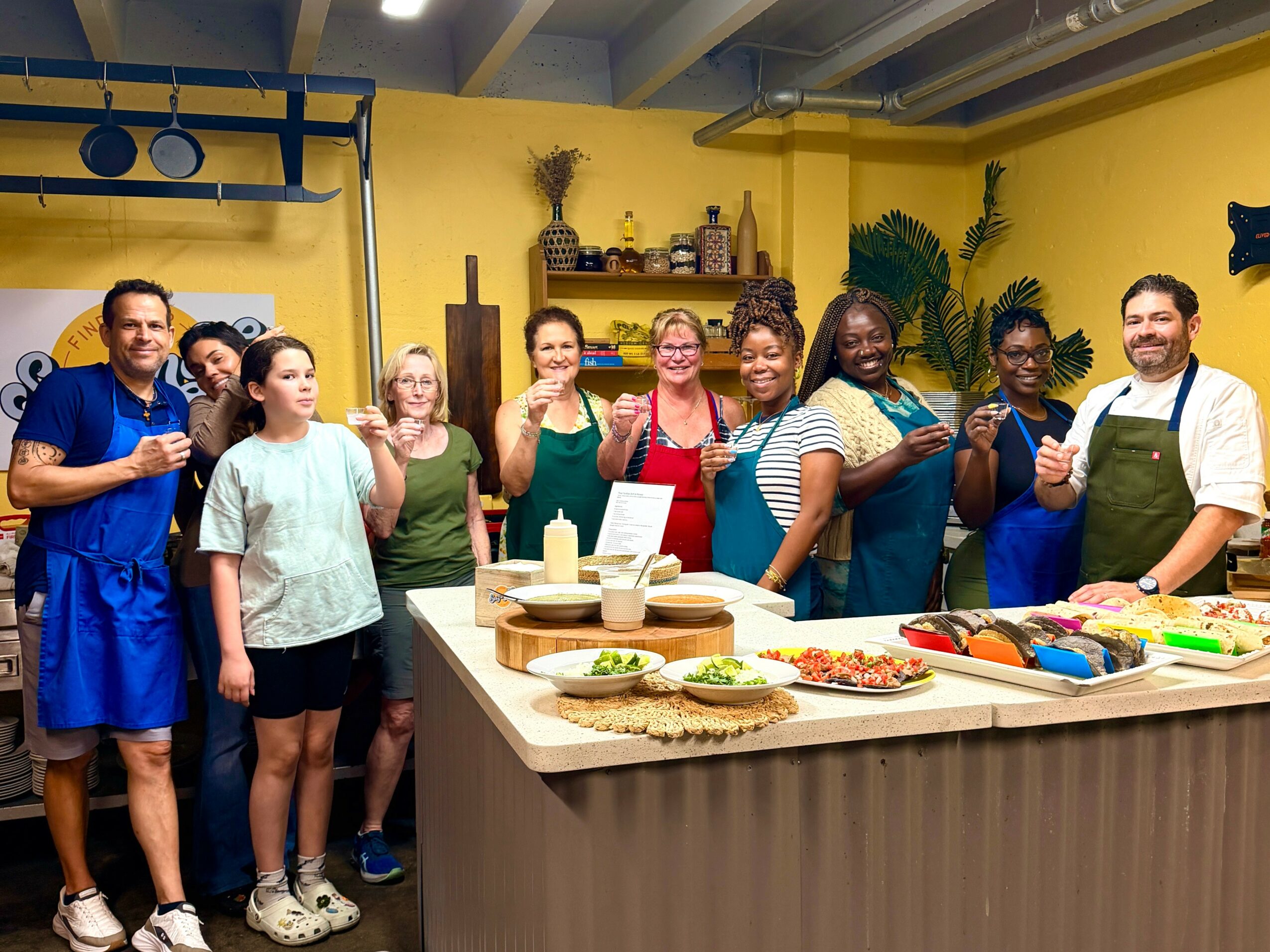 Group of people in a kitchen, smiling and holding drinks, with a counter full of food and drinks, in a yellow room.