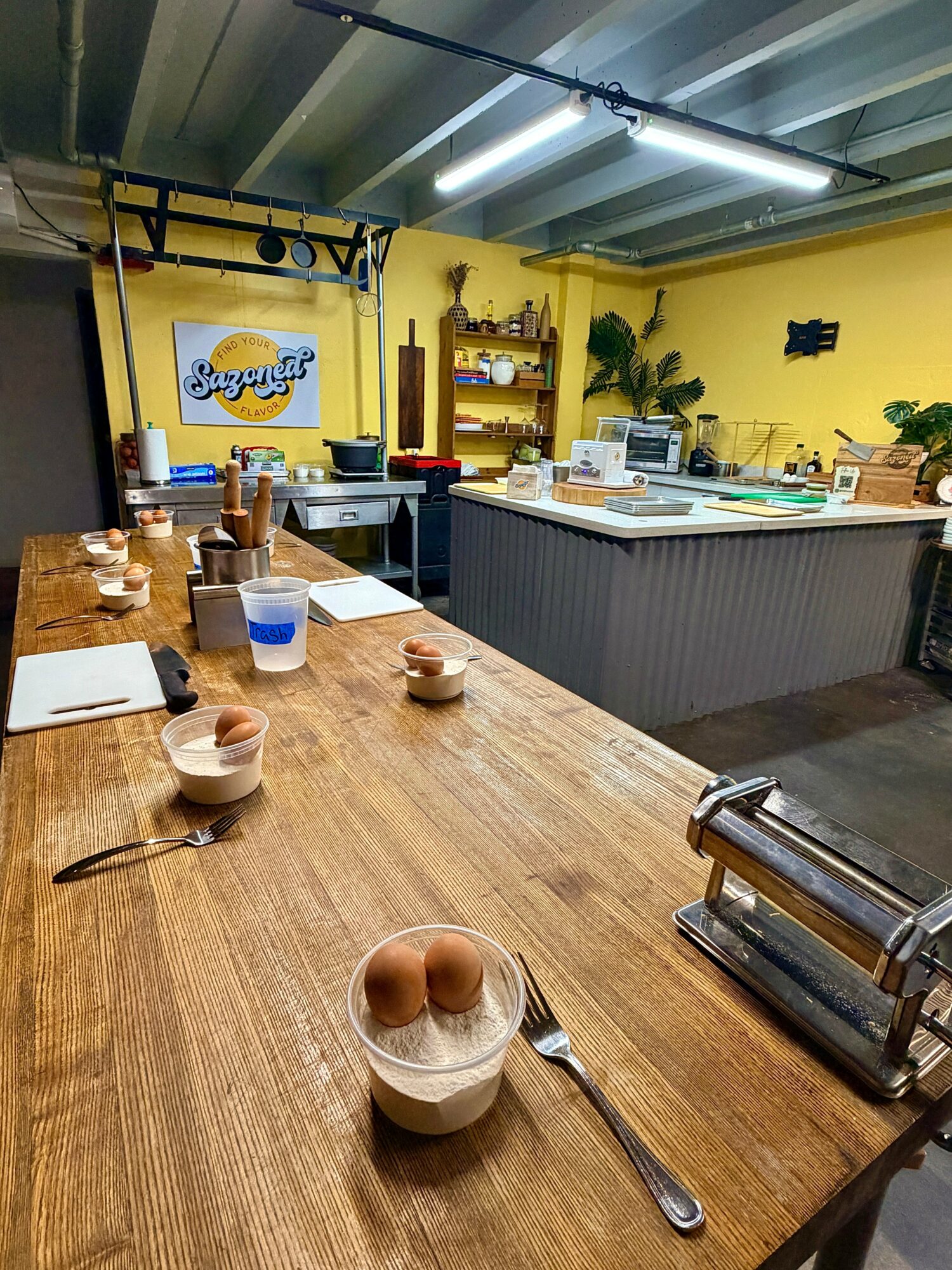 Long wooden counter with bowls, utensils, and glasses, in a yellow-walled cafe with shelves and plants in the background.