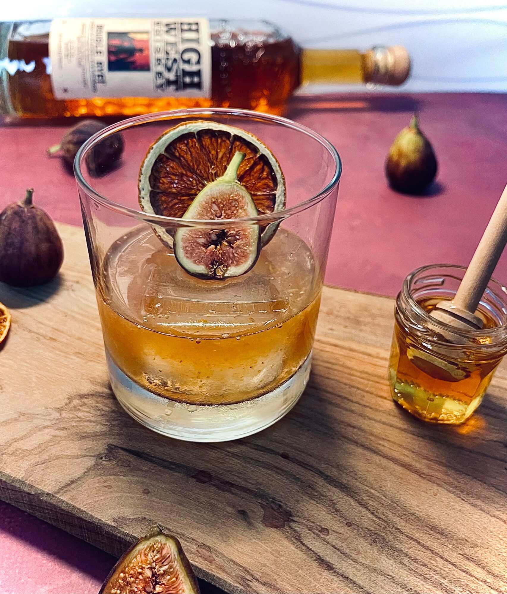 A glass with a drink, garnished with a dried citrus slice, on a wooden surface with honey and figs nearby.