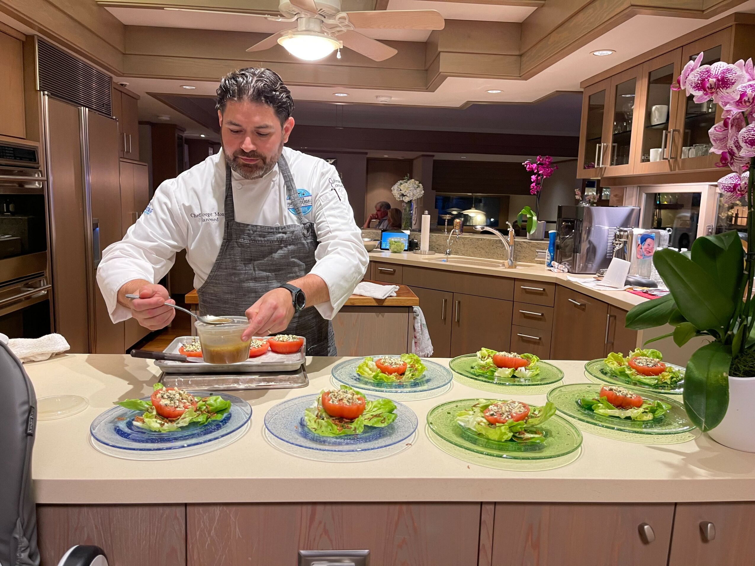 Man in apron preparing food in a kitchen with six plates of salad on the counter, pink flowers in a vase nearby.