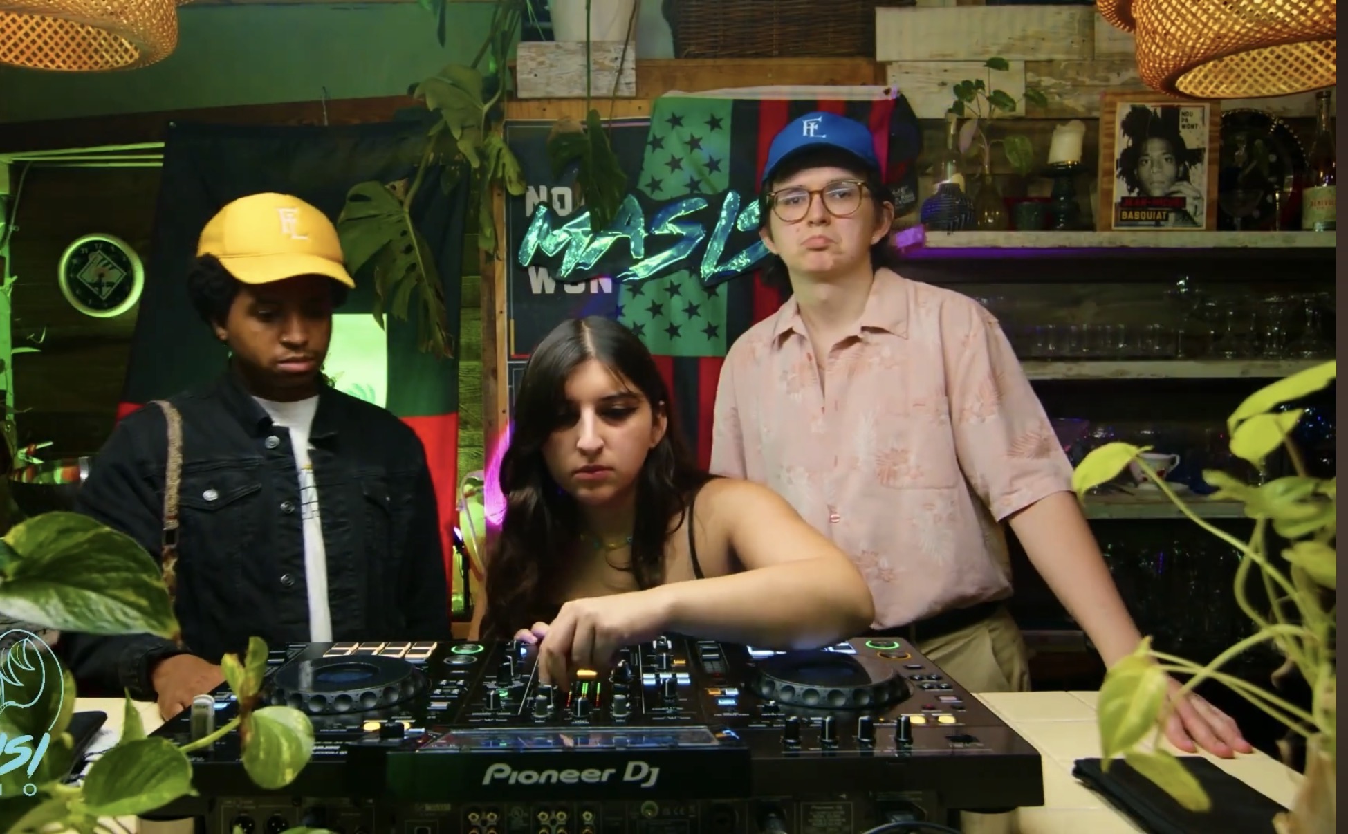 Three people behind a DJ mixer in a colorful indoor setting with plants and decorations.