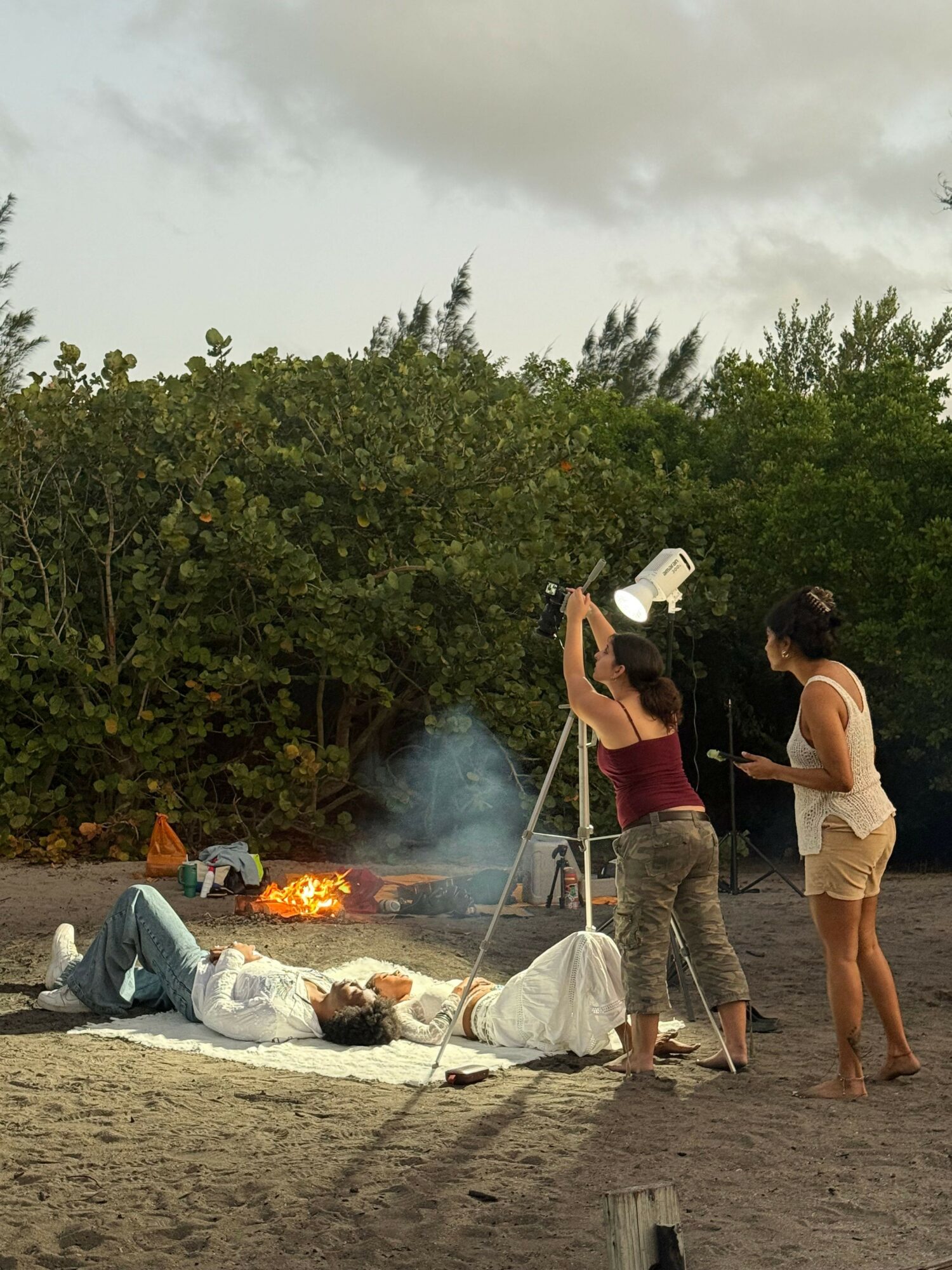 People around a fire outdoors with trees in the background, some standing and some sitting, during daytime.
