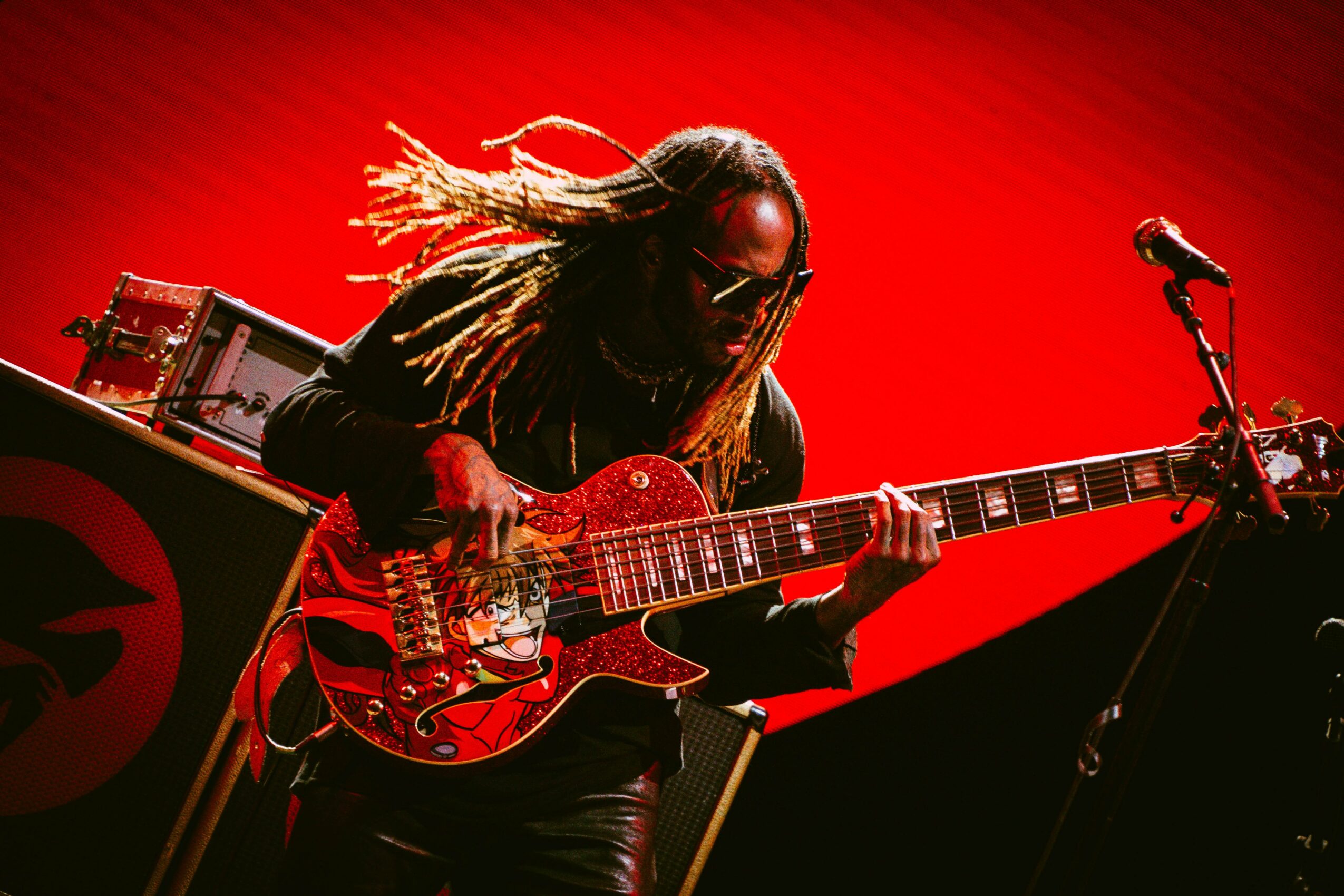 Musician with dreadlocks playing a red electric guitar on stage with a red background.