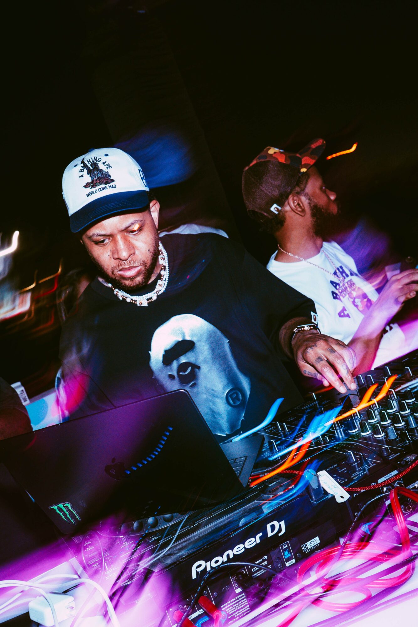 Two DJs perform at a dark club, one wearing a cap and a graphic t-shirt, with colorful lights around them.