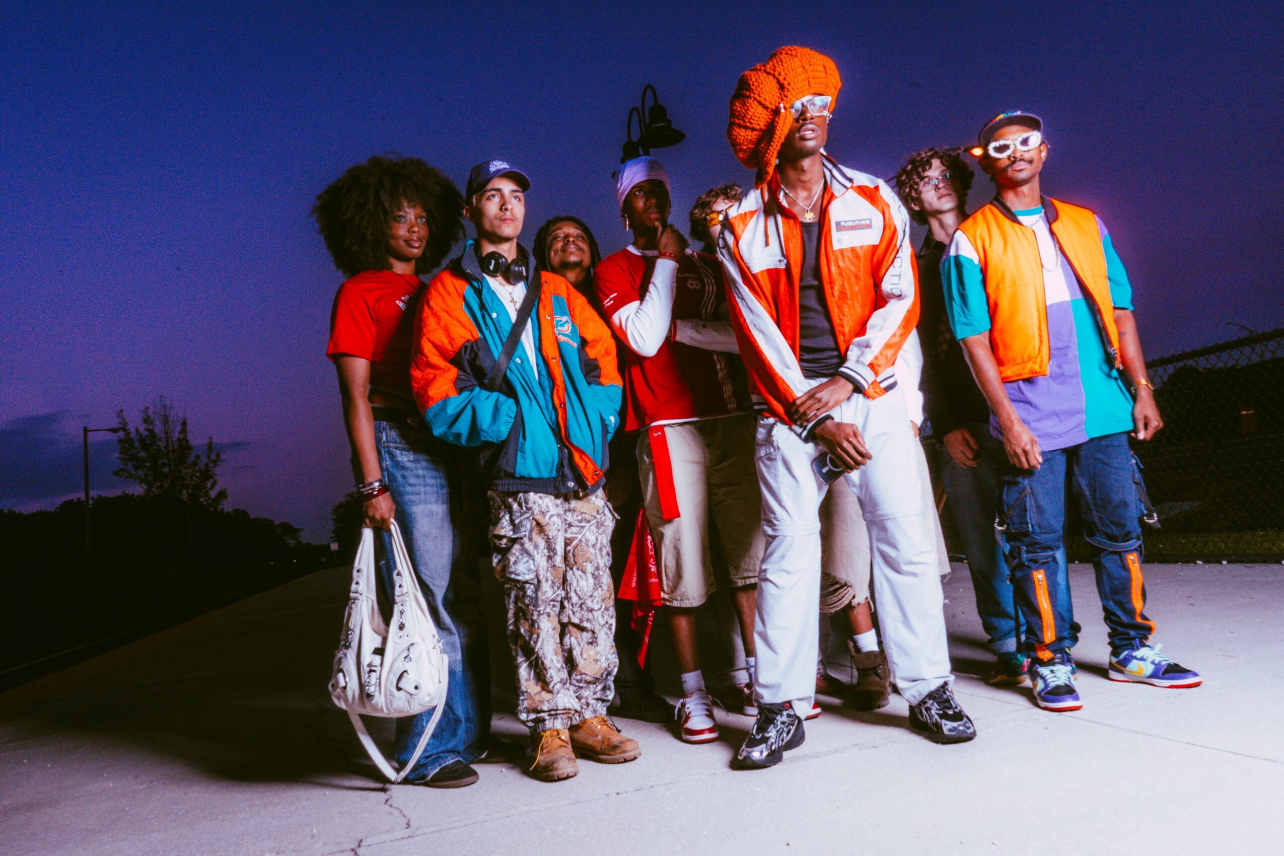 Group of diverse young people standing outdoors at dusk, wearing colorful casual clothing and accessories, some with backpacks.