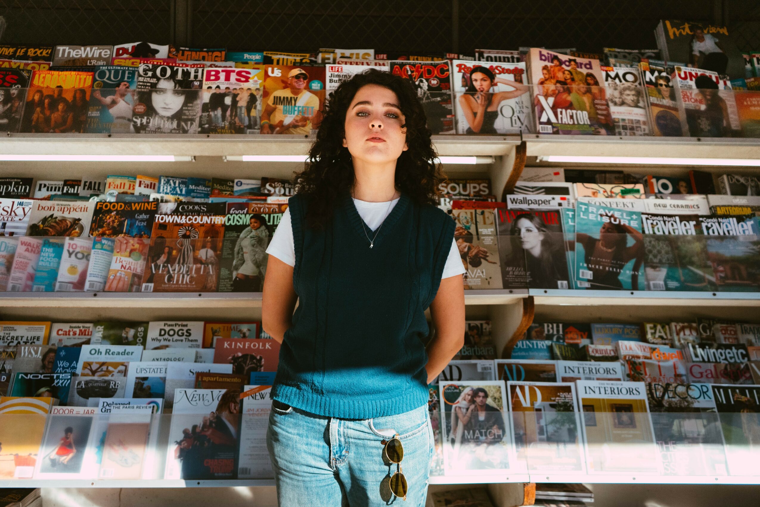 Young woman with dark curly hair stands in front of magazine racks in a store, looking upward. Magazines are displayed behind her.
