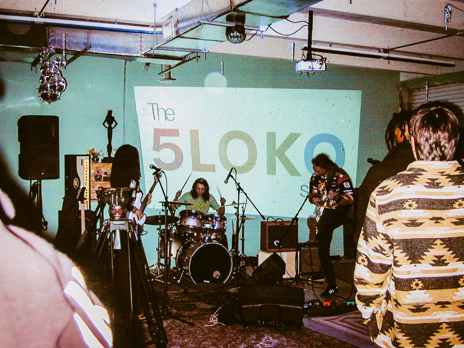 Band performing on stage with a large screen displaying 'The 5LOKO' behind them, audience watching, in an indoor venue.