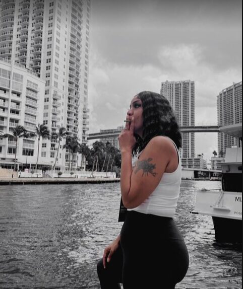 Woman with curly hair and tattoo on arm stands by water in city with tall buildings, bridge, and boat in background.