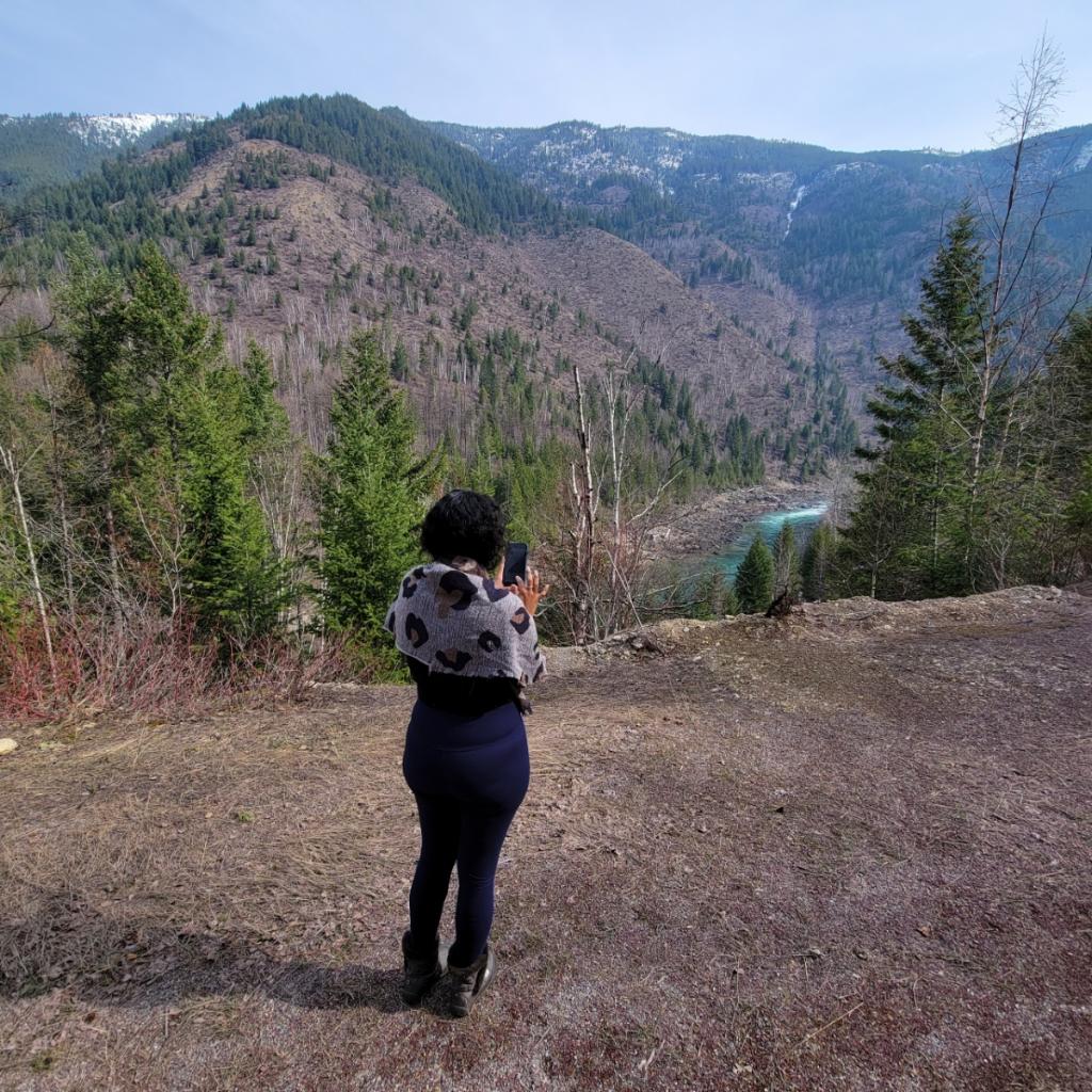 Person standing outdoors on dirt ground, taking a photo of mountains and trees in the distance.