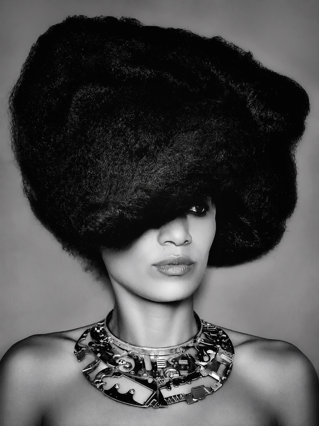 Woman with large, textured hairstyle and bold necklace, looking to the side, in black and white.